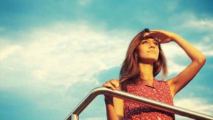 Mulher jovem olhando para o horizonte com a mão na testa para bloquear o sol, apoiada em uma balaustrada, com o céu azul ao fundo, representando a perspectiva e a visão futura que um 'Plano de Carreira: Benefícios para a empresa e colaboradores' pode oferecer.