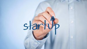 Uma pessoa em uma camisa de botão azul claro escreve a palavra 'startup' no ar com uma caneta, com uma seta apontando para cima substituindo a letra 't', simbolizando crescimento e inovação, elementos chave no contexto de um investidor anjo contribuindo para novos empreendimentos.