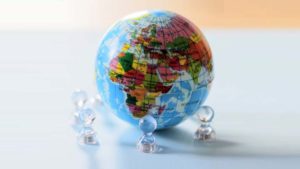 Um globo terrestre colorido, destacando diferentes países em várias cores, está cercado por pequenas figuras transparentes que parecem estar analisando-o, simbolizando a análise global da contabilidade para empresas multinacionais.