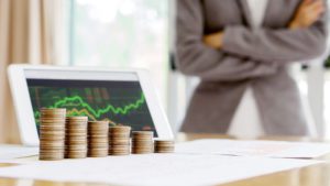 Pilha de moedas crescentes em frente a um tablet mostrando gráficos de desempenho financeiro, com uma pessoa em postura reflexiva ao fundo, para o post 'Corte de custos e despesas: por onde começar'.