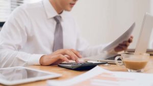 Profissional de camisa e gravata analisando papéis e utilizando uma calculadora em sua mesa de trabalho, com um laptop e uma xícara de café ao lado, representando 'Como calcular o acerto trabalhista'.