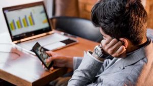 Um homem sentado em uma mesa olha para seu smartphone com uma expressão pensativa, ignorando o laptop aberto com gráficos de barras na tela, ilustrando 'Como começa a procrastinação no trabalho'.