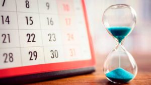 Uma ampulheta com areia azul ao lado de um calendário, elementos que simbolizam a passagem do tempo e prazos, o que pode estar associado ao tema 'Procrastinação no Trabalho', referindo-se ao adiamento de tarefas ou responsabilidades.