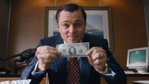 Um homem sorridente segura uma nota de um dólar estendida para a câmera em um escritório bem equipado, evocando a temática do filme "O Lobo de Wall Street", alinhado com o post sobre filmes de contabilidade que exploram o mundo das finanças e da corrupção corporativa.