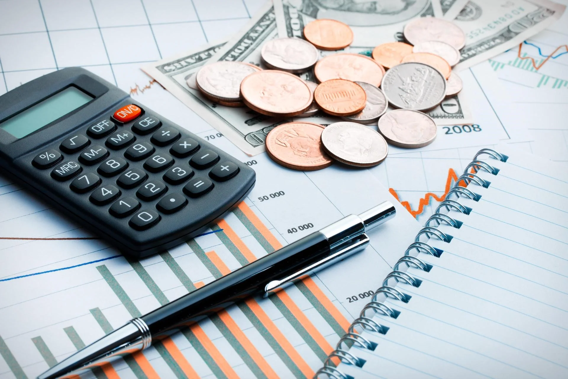 A imagem mostra uma calculadora, moedas, caneta e um bloco de notas sobre gráficos financeiros, elementos que comumente simbolizam a gestão financeira e a otimização fiscal, alinhando-se com o tema '5 formas de Reduzir Impostos da sua Empresa'.