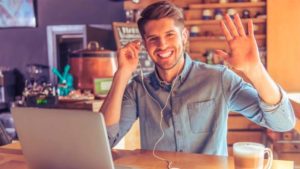 Um homem jovem sorridente acena para a câmera, usando uma camisa azul e fones de ouvido, sentado em frente a um laptop em um café, sugerindo um ambiente de trabalho remoto ou uma videochamada casual.
