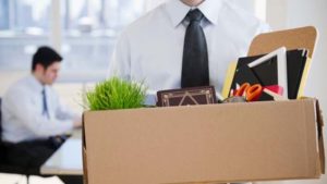 A imagem mostra um indivíduo segurando uma caixa com pertences pessoais de escritório, como uma planta, livros e utensílios, com outra pessoa ao fundo trabalhando, o que alude ao tema de 'Demissão'.