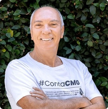Sócio-Fundador Gervásio Souza sorrindo de braços cruzados, vestindo camiseta branca com a hashtag #ContaCMG, em frente a um fundo de folhas verdes.
