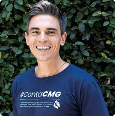 Sócio-Fundador Guilherme Pagotto sorridente, vestindo camiseta azul marinho com a hashtag #ContaCMG, em frente a um fundo de folhas verdes.
