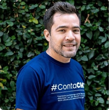 Sócio-Fundador Jonas Oliveira sorridente, vestindo camiseta azul escura com a hashtag #ContaCMG, em frente a um fundo de folhas verdes.