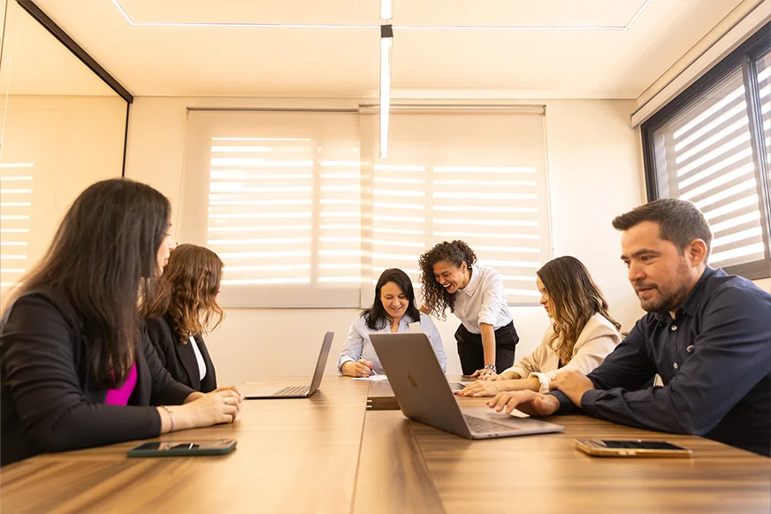Equipe de seis membros da OSP Contabilidade em reunião ao redor de uma mesa com laptops, discutindo ativamente sob a luz natural do escritório.