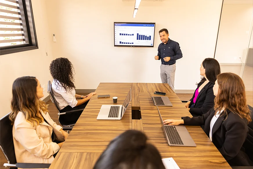Apresentador da OSP Contabilidade de pé mostrando gráficos de barras num monitor para uma equipe de cinco mulheres atentas em uma sala de conferência.