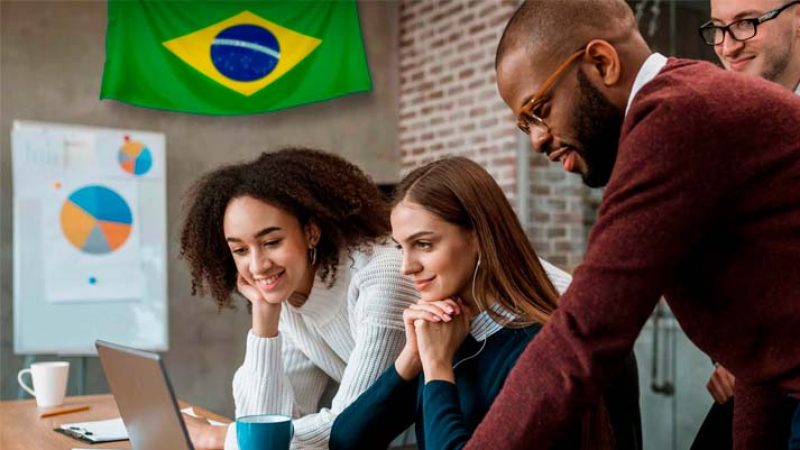 A imagem retrata três profissionais concentrados olhando para um laptop, com um gráfico circular ao fundo e uma bandeira do Brasil pendurada na parede, evocando temas de diversidade e colaboração internacional no contexto de 'Contabilidade para Multinacionais'.